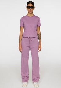 Lilla to-delte sæt bestående af en T-shirt med korte ærmer og bukser med snoretræk, lavet af blødt stof, parret med mørke solbriller og hvide sneakers.
