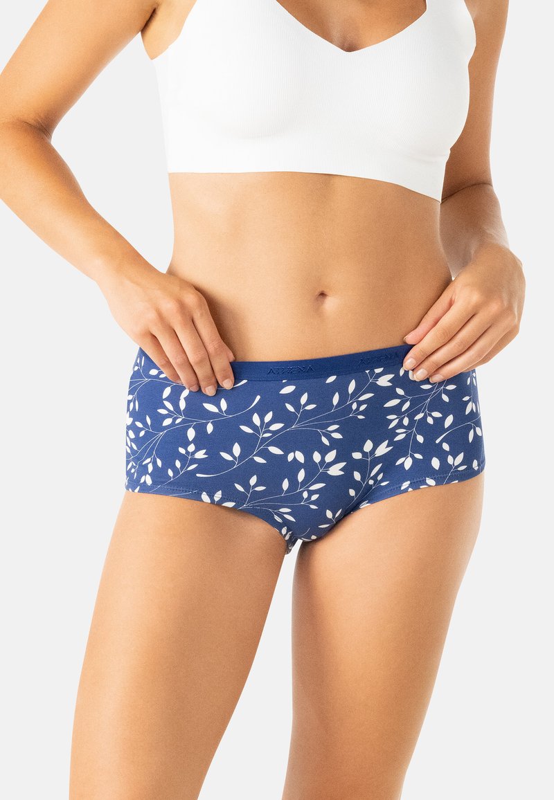 Femme portant un soutien-gorge de sport blanc et des sous-vêtements bleus avec un motif de feuilles blanches, tenant la taille des sous-vêtements.