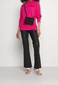 Persona che indossa una felpa fucsia brillante, pantaloni in pelle nera, sandali con tacco bianchi e una borsa a tracolla nera su uno sfondo semplice.