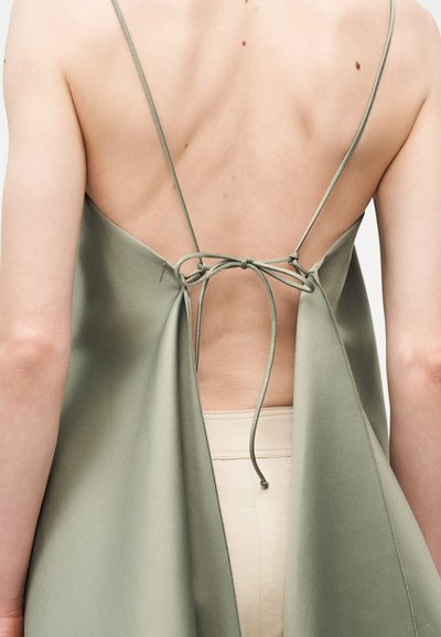 Άτομο που φοράει ένα φόρεμα ανοιχτής πλάτης σε φιστικί πράσινο χρώμα, δεμένο με λεπτές τιράντες, αποκαλύπτοντας το άνω μέρος της πλάτης και τη ζώνη ενός μπεζ εσωρουχου.