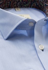 Lichtblauw overhemd voor heren met witte knopen, klassieke kraag en een patroon aan de binnenkant van de kraag.