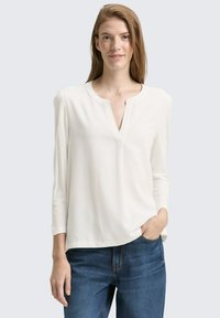 Blusa bianca a maniche lunghe con scollo a placket, texture liscia e vestibilità rilassata, abbinata a jeans in denim blu.