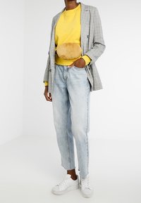 Osoba v žluté mikině, šedém kostkovaném blejzru, světle modrých džínách, bílých teniskách a béžové huňaté ledvince přes pas.