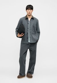 Uomo con giacca e pantaloni in denim grigio che tiene una borsa nera, indossa scarpe marroni, in piedi contro uno sfondo bianco semplice.