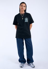 Camiseta de algodón negra con un diseño gráfico en azul claro en el lado izquierdo del pecho. Combinada con jeans de pierna ancha azules y zapato deportivos plateados.