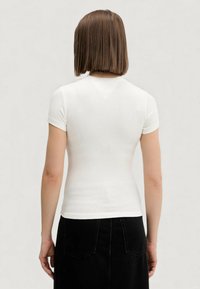 Bílá žebrovaná tričko s krátkým rukávem a přiléhavým střihem, se zády ve tvaru V a hladkou texturou. Nošeno s černou sukní.