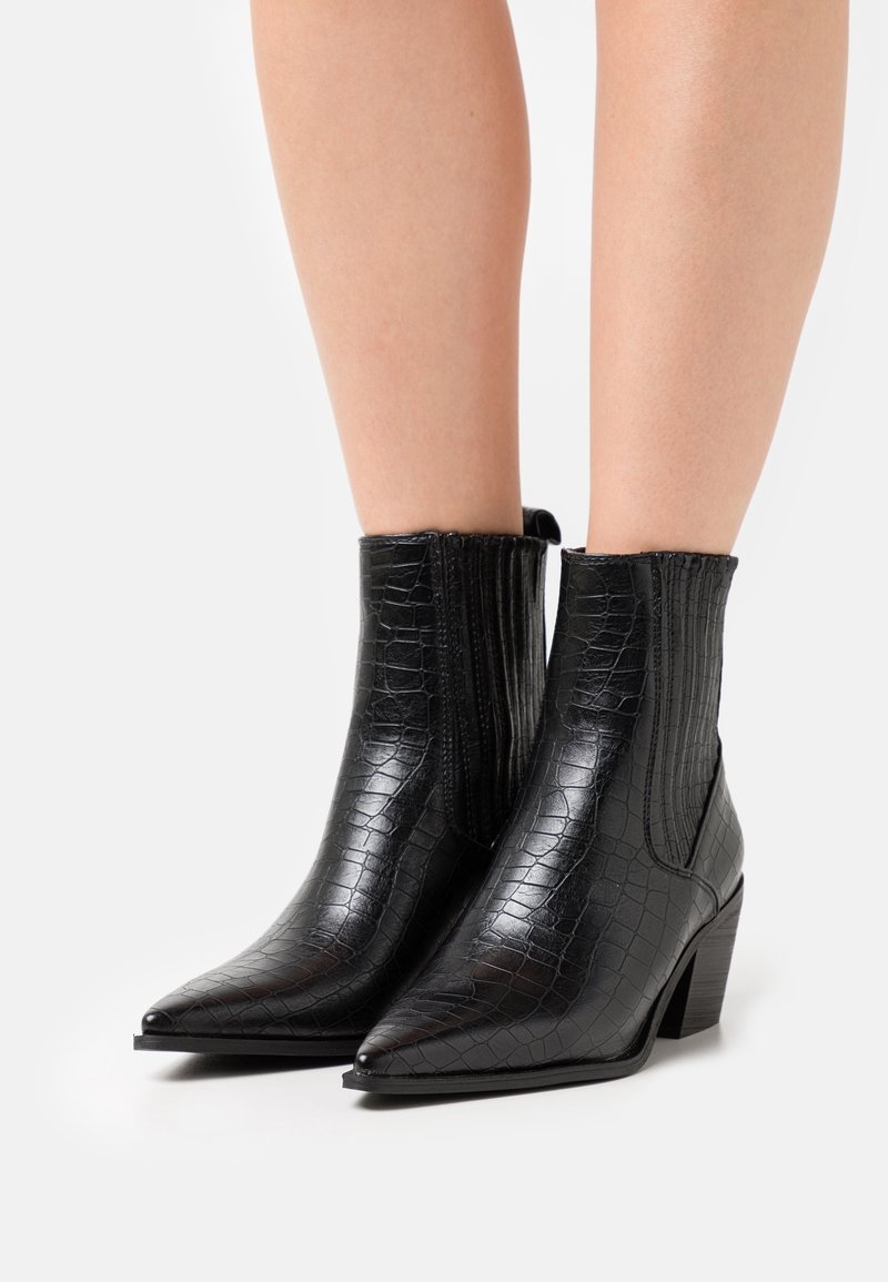 Bottes cheville à bout pointu en cuir noir à texture de crocodile avec talons épais, portées sur des jambes nues sur fond blanc.