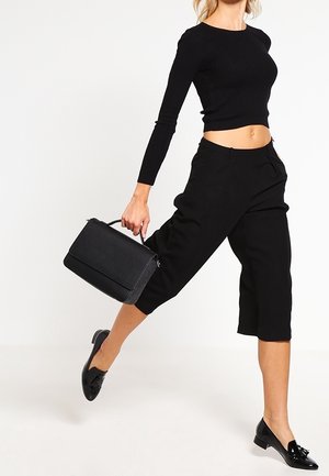 Bolso estructurado negro con un asa superior, textura suave y cierre con cremallera. Llevado con una blusa negra de canalé ajustada y pantalones cortos.