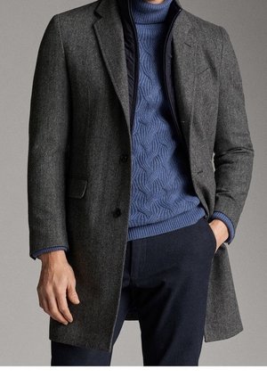 Homme portant un manteau en laine gris sur un pull texturé bleu et un pantalon sombre, une main dans la poche et l'autre le long du corps.