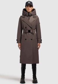 Trench coat lung, maro, cu glugă, cu un front îndoit, talie cu cingătoare și butoane mari, asortat cu cizme de luptă negre și o pălărie.