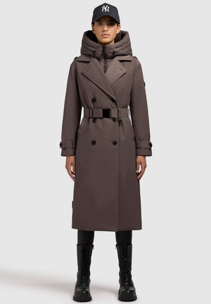 Langer brauner Trenchcoat mit Kapuze, doppelreihiger Front, tailliertem Gürtel und großen Knöpfen, kombiniert mit schwarzen Stiefeln und einer Mütze.