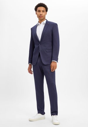 Young man standing wearing a navy blue suit with a white shirt and white sneakers against a plain white background.