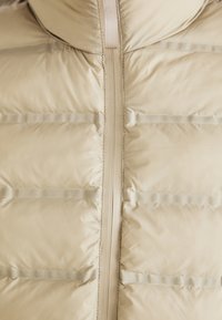 Chaqueta acolchada beige con una textura suave, que presenta secciones acolchadas horizontales y una cremallera central. Diseño minimalista sin adornos.