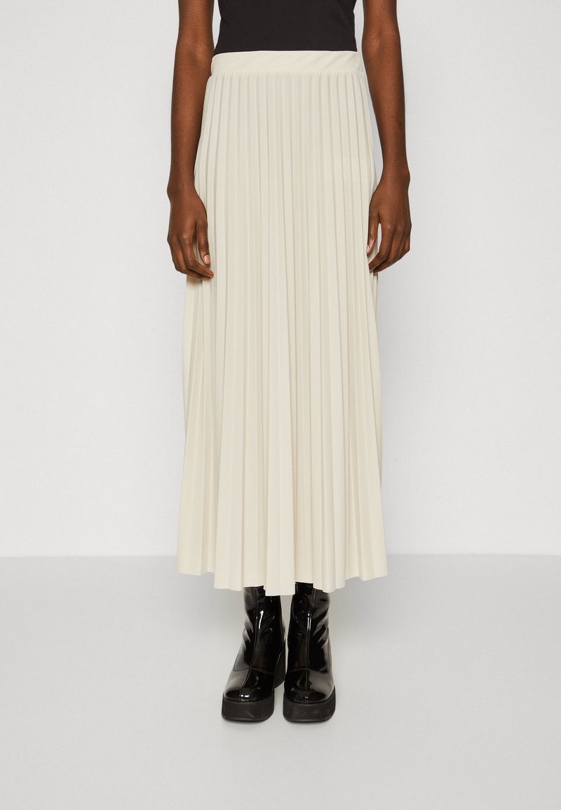 Femme portant une jupe midi plissée crème avec un top noir ajusté et des bottines à plateforme noires brillantes, debout devant un fond uni.