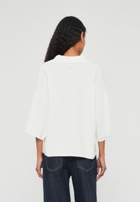 Top de malha oversized branco com colarinho e mangas curtas, apresentando um acabamento texturizado e um discreto detalhe de logotipo nas costas. Parte inferior em ganga escura.
