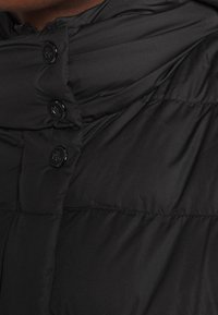 Musta toppatakki, jossa on korkea kaulus, kiiltävät mustat napit ja tikattu pinta, korostettuna hienovaraisilla saumoilla ja taitoksilla.