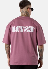 Mann mit Nackentattoo und Sonnenbrille, der ein oversized T-Shirt in Mauve trägt, das auf der Rückseite weißen grafischen Text zeigt, während seine tätowierten Arme sichtbar sind.