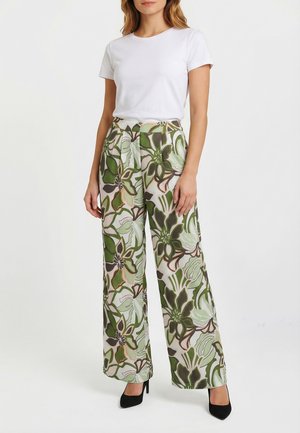 Femme portant une chemise blanche à manches courtes, un pantalon large taille haute à grand motif floral vert, et des talons noirs, debout sur un fond blanc.