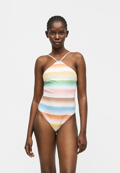 Femme portant un maillot de bain une pièce à rayures multicolores et à col haut, avec de fines bretelles, debout devant un fond clair uni.