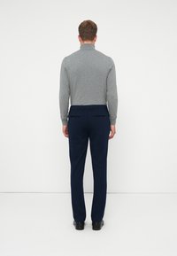 Muž stojí čelom k bielej stene, oblečený v šedých rolákoch, námorníckych nohaviciach s zadnými vreckami a čiernych formálnych topánkach.