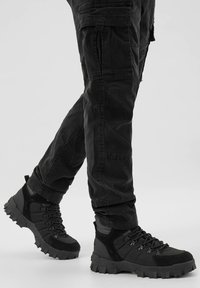 Černé termoskové boty na turistiku vyrobené z kombinace texturovaných materiálů. Mají robustní podešve, šněrování a polstrovaný kotník pro podporu.