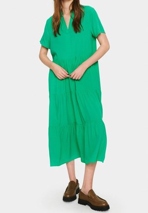 Femme portant une robe midi verte ample à pans superposés et des mocassins marron à semelle épaisse, debout, mains jointes à la taille sur un fond blanc uni.