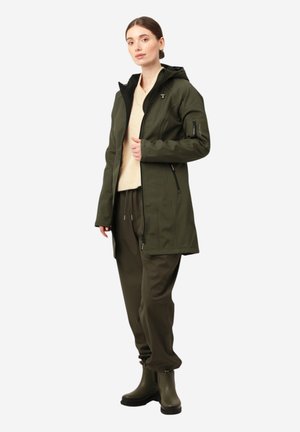 Chaqueta impermeable verde oliva con capucha, bolsillos con cremallera y detalle de cremallera en el lateral, llevada sobre un suéter crema y pantalones oliva.