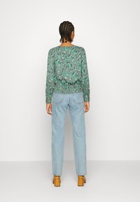 Camisa de manga longa verde com estampa floral e bainha franzida, combinada com jeans de cintura alta azul claro e sapatos de salto bloco em tom beige.