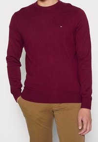 Bordó pletený svetr s kulatým výstřihem a žebrovanými manžetami. Malé logo v červené, bílé a modré barvě na prsou. Měkká textura.