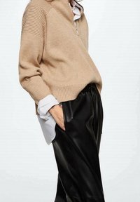 Beżowy sweter z dzianiny z kołnierzem na zamek, z teksturowanymi rękawami, nałożony na białą koszulę. Połączony z czarnymi skórzanymi spodniami o wysokim stanie.