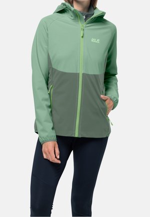 Persona vistiendo una chaqueta con capucha de dos tonos de verde de Jack Wolfskin con cremallera y bolsillos, combinada con pantalones negros, de pie contra un fondo liso.