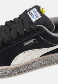 Schwarzer Wildleder-Sneaker mit weißen, rissigen Lederakzenten, strukturiertem grauem Außenlauf, schwarzen Schnürsenkeln und einem bunten Markenetikett nahe der Zunge.