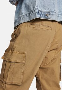 Beige cargo pants with side pockets and textured fabric; paired with a light blue denim jacket featuring a button detail.