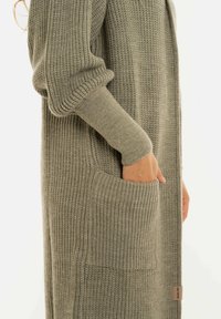 Zijaanzicht van een persoon in een lange, olijfgroene gebreide cardigan met de hand in de voorzak en losse mouwen.