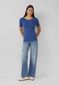 Kobieta z długimi brązowymi włosami, ubrana w niebieską koszulę z krótkim rękawem, jasnoniebieskie szerokie dżinsy oraz białe trampki, stoi przy szarej ścianie.