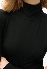 Eine Frau trägt ein schlichtes schwarzes Langarm-Turtleneck-Oberteil und hat schulterlanges, hellbraunes Haar vor einem weißen Hintergrund.