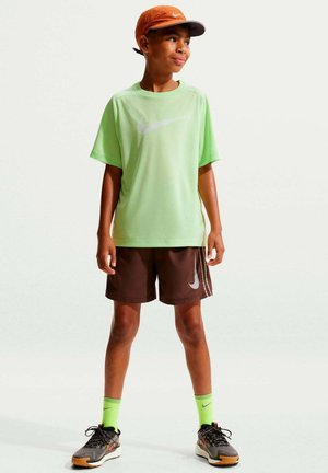 Niño con camiseta Nike verde lima, pantalones cortos Nike marrones, calcetines Nike verde neón, zapatillas grises y naranjas y gorra naranja, de pie mirando hacia la derecha.
