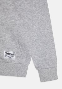 Sivi pamučni dukserica s kenguruvim džepom, rebrastim manžetama i pravokutnom Timberland oznakom na donjoj strani. Mekana tekstura.