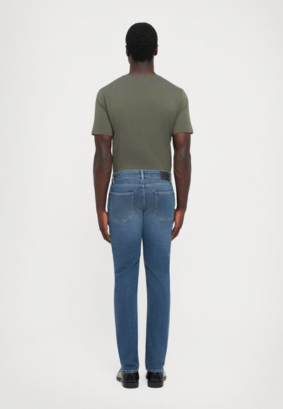 Man standing facing away wearing an olive green t-shirt, blue jeans with back pockets, and black shoes against a plain white background.