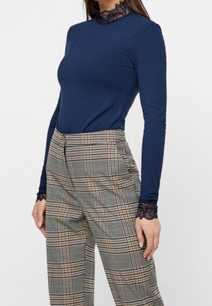 Mujer con un top de manga larga azul marino con cuello y puños de encaje, combinado con pantalones de tiro alto a cuadros en tonos beige y negro.