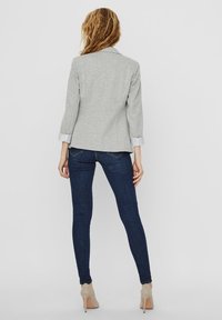 Blazer gris avec une coupe ajustée et des manches trois-quarts, associé à un jean skinny bleu foncé et des escarpins beiges. Tissu texturé et poignets contrastés.