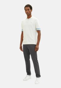 Weißes Poloshirt mit blau gestreiften Ärmeln, kombiniert mit dunkelgrauen Hosen und weißen Sneakers. Kurze Ärmel, normale Passform und minimales Branding.