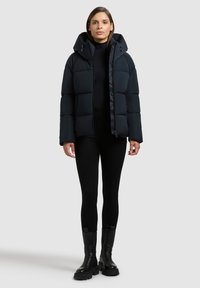 Giubbino nero invernale con cappuccio, caratterizzato da un design trapuntato e tasche laterali, abbinato a legging neri aderenti e stivali neri robusti.