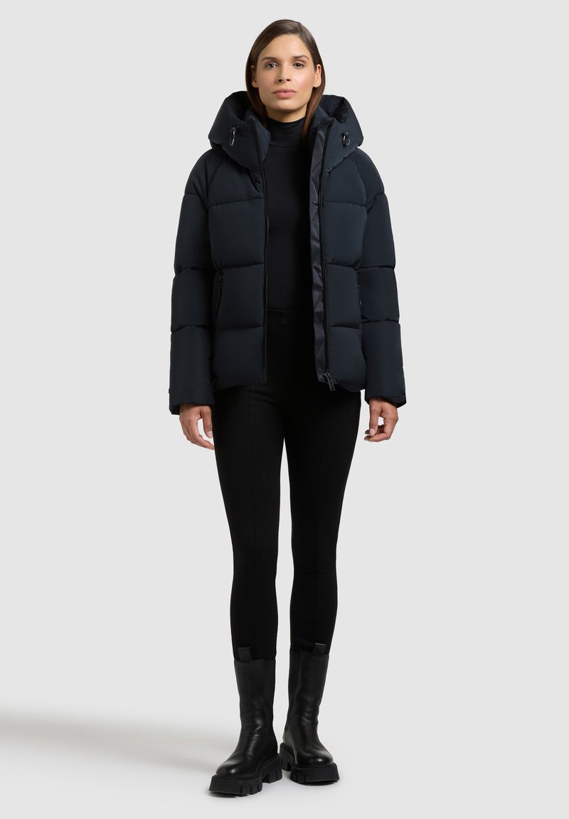 Giubbino nero invernale con cappuccio, caratterizzato da un design trapuntato e tasche laterali, abbinato a legging neri aderenti e stivali neri robusti.