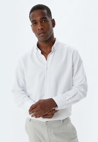 Camisa blanca de manga larga con cuello, textura suave, y puños con botones, combinada con pantalones grises claros.