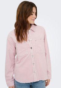 Jonge vrouw draagt een lichtroze corduroy blouse met knopen, twee borstzakken en een rafelige zoom, gecombineerd met een blauwe spijkerbroek.