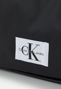 Svart tygväska med en vit etikett som har "Calvin Klein Jeans" och en stiliserad "CK"-logotyp. Slät textur med sydda kanter.