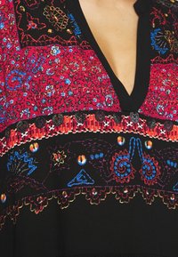 Robe noire à décolleté en V profond, ornée de motifs complexes de broderies florales et géométriques rouges, bleues et orange sur la poitrine et la taille.