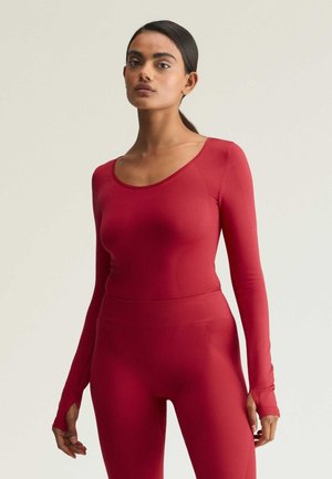 Vrouw met donker haar in een lage paardenstaart, gekleed in een strakke rode top met lange mouwen en bijpassende rode legging, staand voor een effen lichte achtergrond.
