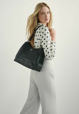 Blondynka w białych spodniach i białej bluzce w czarne groszki, niosąca dużą czarną skórzaną torebkę na ramieniu.